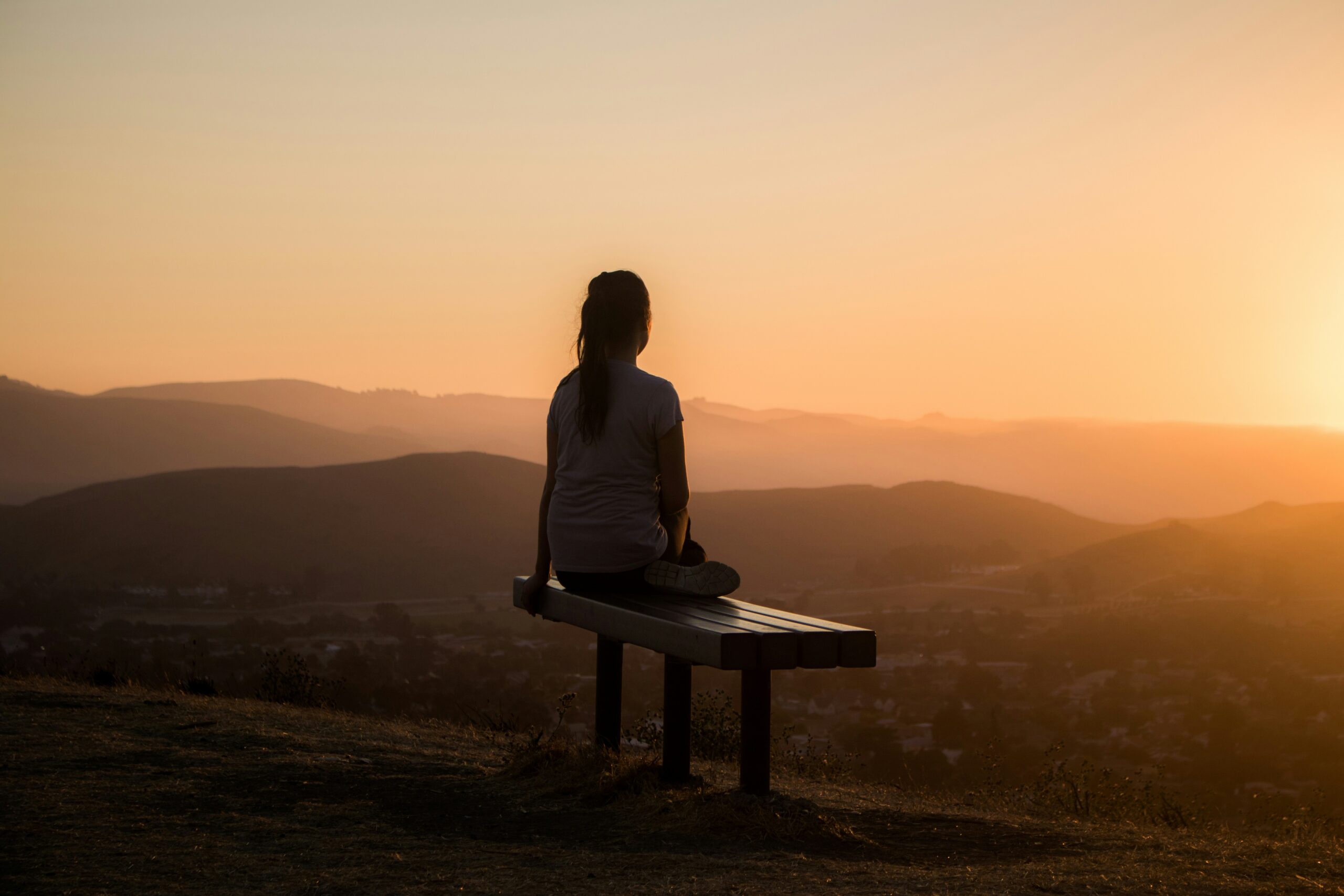 person sitting crosslegged on bench at sunset with scenic mountain view